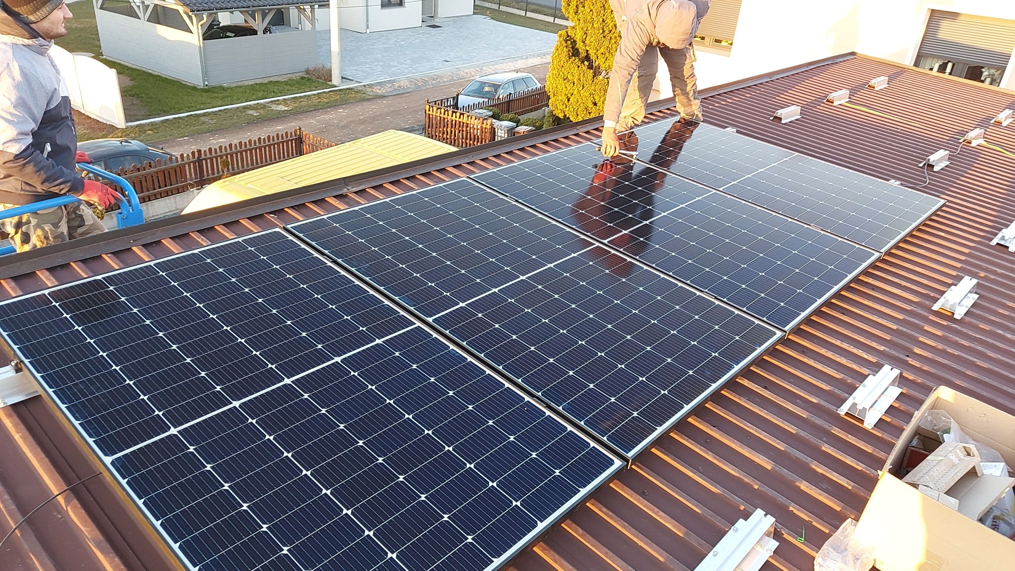 Montaż paneli słonecznych na brązowym dachu z blachy trapezowej, widoczne elementy montażowe i pracownicy w trakcie instalacji.