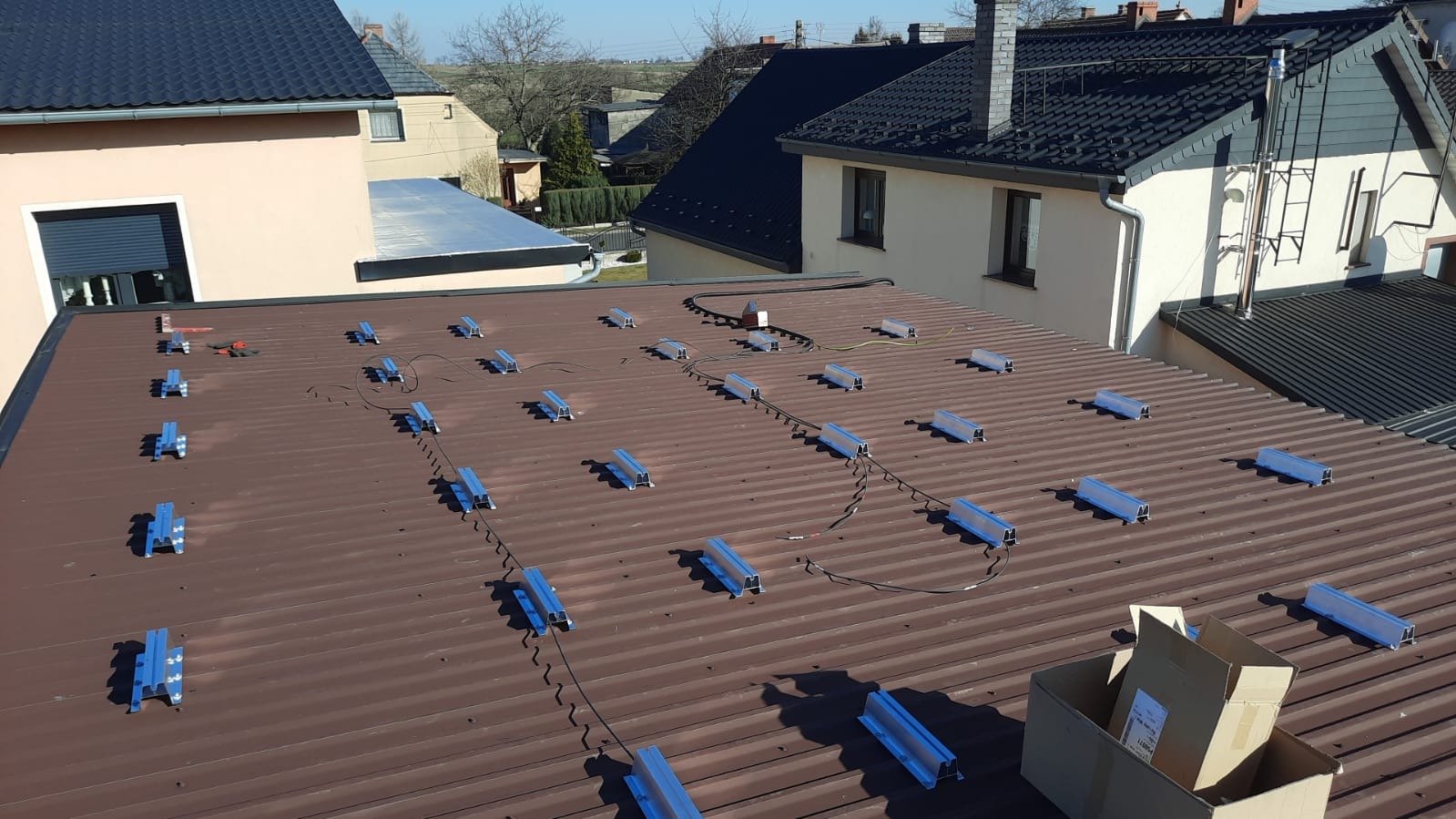 Brązowy dach z zamontowanymi niebieskimi uchwytami montażowymi pod panele słoneczne, widoczne kable i kartonowe pudełko z elementami, w tle zabudowa jednorodzinna.
