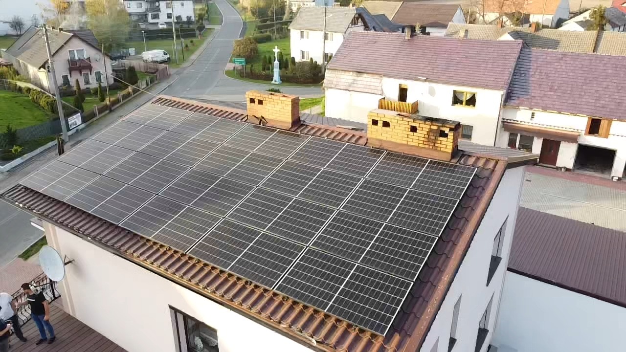 Widok z góry na panele słoneczne na dachu domu jednorodzinnego z ceglanymi kominami, w tle zabudowa wiejska i droga.