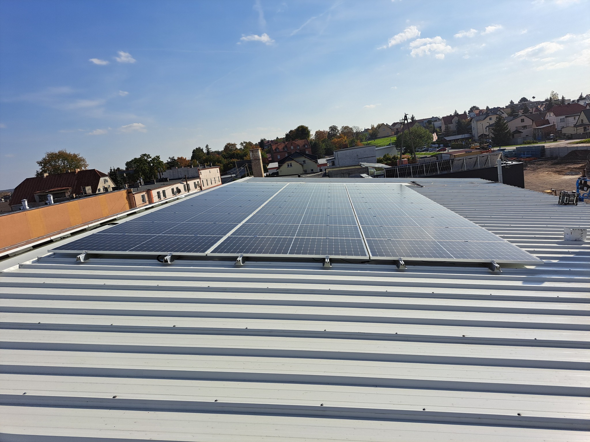 Panele słoneczne zamontowane na blaszanym dachu budynku, widok z góry na tle zabudowań miejskich i błękitnego nieba.
