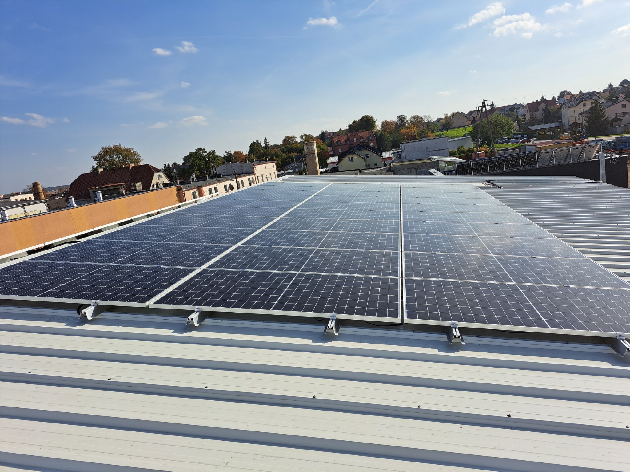 Rząd paneli słonecznych na blaszanym dachu budynku w słoneczny dzień, widok na miasto w tle.