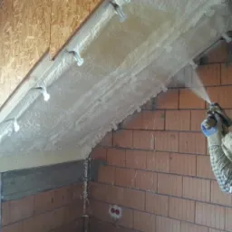 Aplikacja pianki izolacyjnej na poddaszu z widocznymi dyblami mocującymi, operator w rękawicach ochronnych spryskuje powierzchnię, ceglana ściana w tle.
