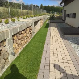 Nowoczesny ogród z równo przystrzyżonym trawnikiem, kamienną ścieżką, murem z miejscem na drewno opałowe i ozdobnymi krzewami wzdłuż ogrodzenia.