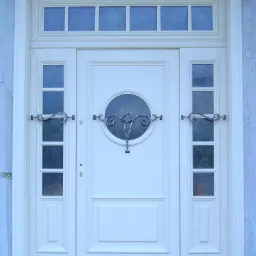 Eleganckie białe drzwi wejściowe z okrągłym przeszkleniem i ozdobnymi metalowymi uchwytami, otoczone bocznymi panelami z szybkami i górnym świetlikiem, osadzone w klasycznej ramie.