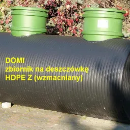 Czarny, karbowany zbiornik na deszczówkę z dwoma zielonymi pokrywami inspekcyjnymi, umieszczony na kostce brukowej przed ogrodem z czerwonymi liśćmi.