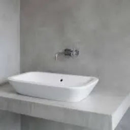 Minimalistyczna łazienka z białą wanną wolnostojącą na betonowym blacie, surowe szare ściany z widoczną fakturą betonu, chromowana bateria ścienna.