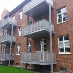 Budynek z czerwonej cegły z metalowymi balkonami na konstrukcji stalowej, widok z dołu na zewnątrz, trawnik przed budynkiem.