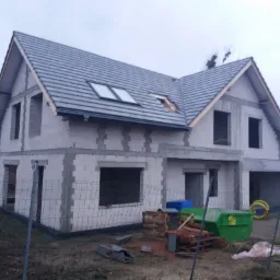 Surowy stan dwukondygnacyjnego domu z szarej cegły z szarym dachem, widoczne okna dachowe i otwory okienne, otoczone tymczasowym ogrodzeniem z siatki, z paletami i pojemnikami na gruz na pierwszym...