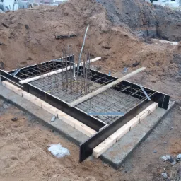 Zbrojenie i szalunek pod fundament w wykopie, widoczne pręty pionowe i poziome, drewniane podpory i czarna płyta szalunkowa.