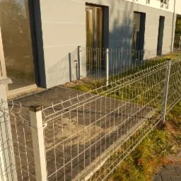 Nowoczesne, metalowe ogrodzenie panelowe w kolorze srebrnym, otaczające posesję z kostką brukową i budynkiem w tle. Widok z perspektywy ziemi, słoneczny dzień.