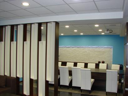 Nowoczesny wystrój biura: dekoracyjna ścianka działowa z drewnianymi elementami, stół konferencyjny z białymi krzesłami, oświetlenie sufitowe.