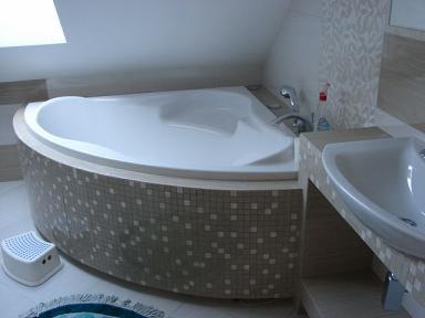 Nowoczesna, narożna wanna obłożona mozaiką w jasnej łazience na poddaszu, z widoczną umywalką i dziecięcym stołkiem. Ujęcie z niskiego kąta.