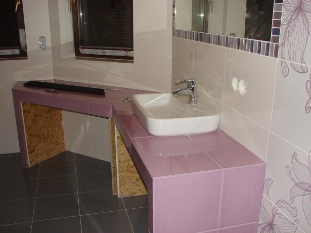 Łazienka w trakcie remontu. Umywalka nablatowa osadzona na różowym blacie z płytek, częściowo wykończone ściany z motywem kwiatowym, widoczne okna i szara podłoga.