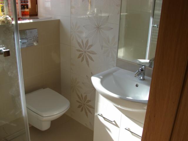 Jasna łazienka z umywalką na szafce, toaletą i kabiną prysznicową. Ściany wykończone płytkami z delikatnym, kwiatowym wzorem. Ujęcie z perspektywy wejścia do kabiny.