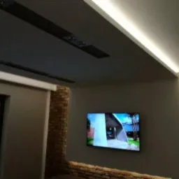 Nowoczesne wnętrze z podwieszanym sufitem w kolorze grafitowym, oświetleniem LED wzdłuż krawędzi sufitu, ścianą z cegły dekoracyjnej i telewizorem LCD.