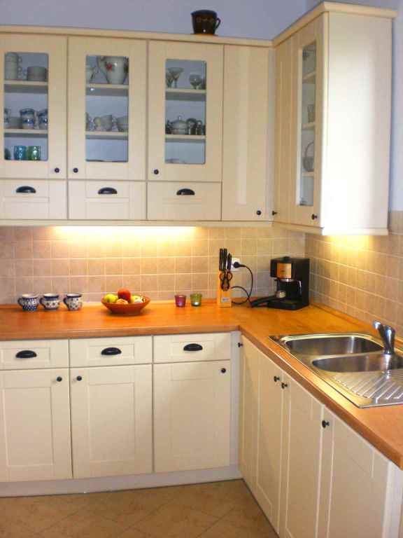 Jasna kuchnia z drewnianym blatem, szafki górne z przeszklonymi frontami, na blacie owoce, filiżanki i ekspres do kawy.