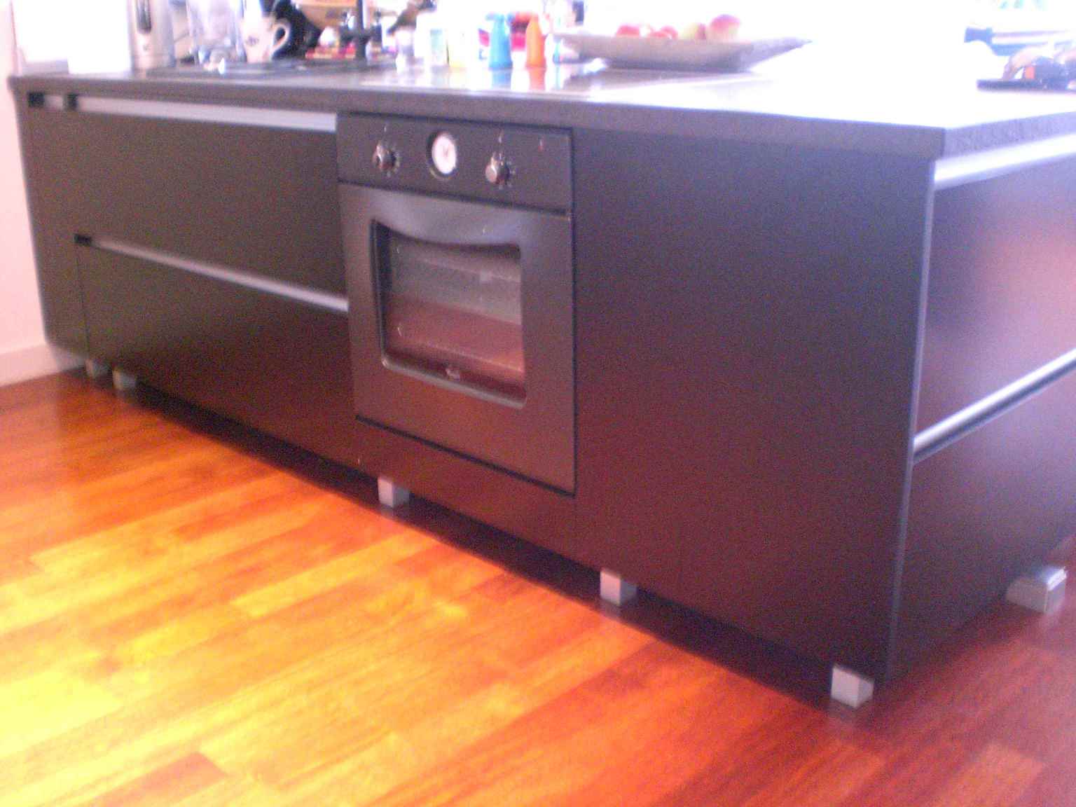 Ciemna zabudowa kuchenna z piekarnikiem wbudowanym w szafkę, widoczna z niskiego kąta na tle parkietu. Minimalistyczny design z prostymi liniami i metalowymi uchwytami.