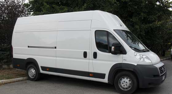 Biały, dostawczy Fiat Ducato zaparkowany na tle zielonych drzew, z bocznymi drzwiami przesuwnymi i relingiem dachowym.