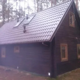 Ciemnobrązowy dom z bali z metalowym dachem w lesie. Widoczne okna z drewnianymi ramami, rynna i kominy. Dach pokryty brązową blachodachówką z oknami dachowymi.