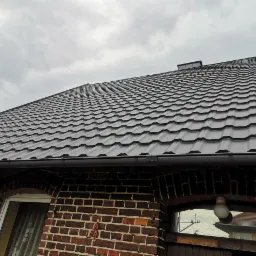 Nowy, ciemnoszary dach z blachodachówki modułowej na budynku z czerwonej cegły, widok z dołu w pochmurny dzień.