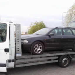 Czarny samoch&oacute;d kombi marki Audi A4 z otwartą klapą bagażnika, przewożony na platformie białej lawety w pochmurny dzień.
