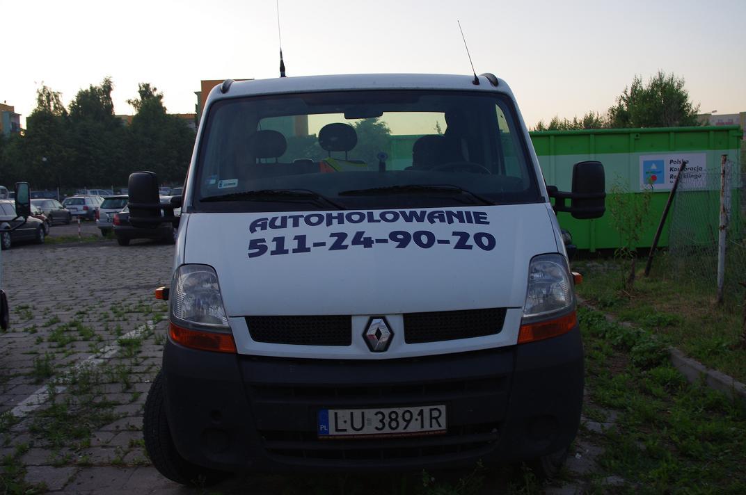 Biały bus Renault z napisem 'AUTOHOLOWANIE 511-24-90-20' na przedniej masce, zaparkowany na placu z kostki brukowej, w tle zielony kontener i inne zaparkowane samochody.