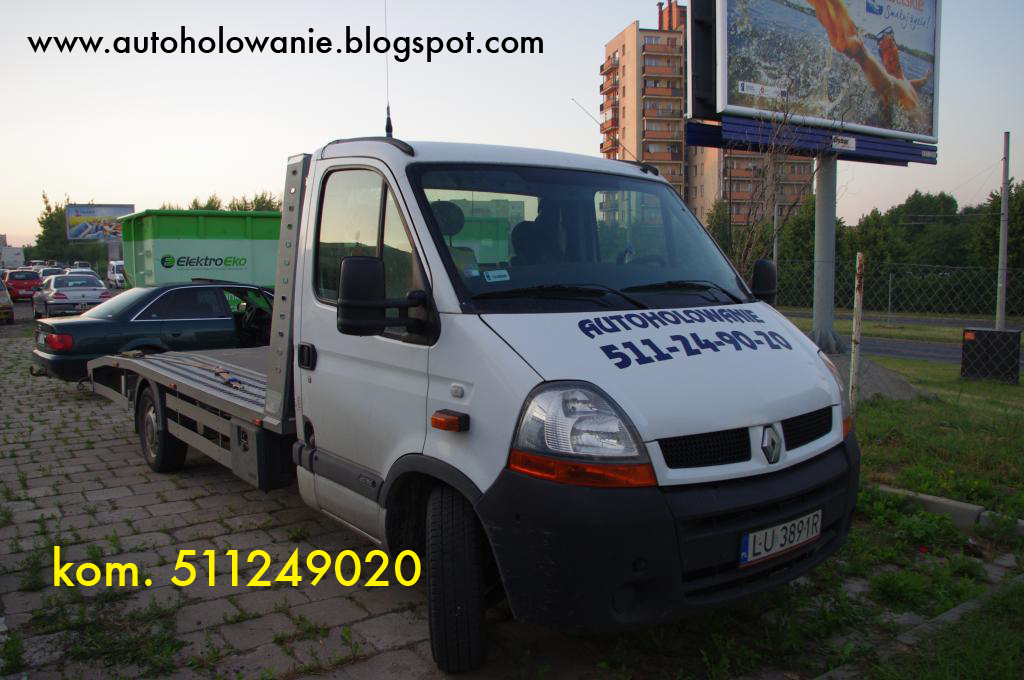 Biała laweta Renault Master z napisem 'AUTOHOLOWANIE 511-24-90-20' na przedniej szybie, zaparkowana na brukowanej powierzchni w pobliżu zielonego kontenera ElektroEko i billboardu reklamowego.