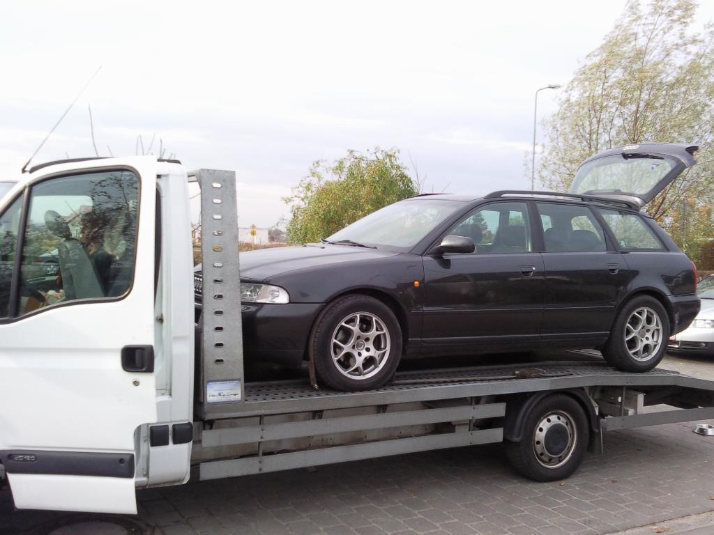 Czarny samochód kombi marki Audi A4 z otwartą klapą bagażnika, przewożony na platformie białej lawety w pochmurny dzień.