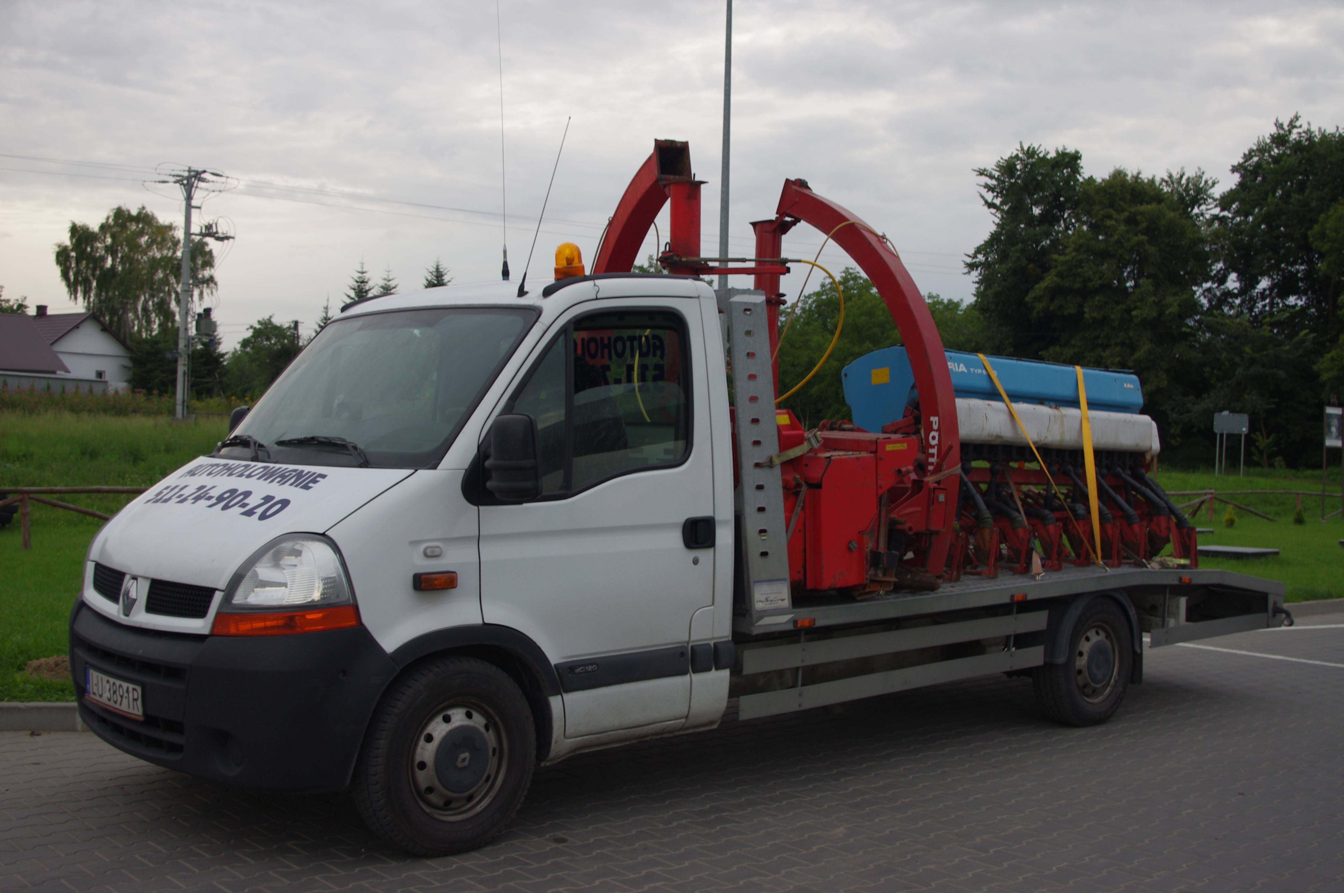Biały bus z platformą transportową przewożący czerwoną maszynę rolniczą z niebieskim zbiornikiem, zabezpieczoną żółtymi pasami.