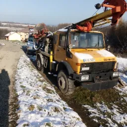 Żółta ciężarówka z wiertnicą do studni głębinowych zaparkowana na poboczu drogi w zimowym krajobrazie, widoczny numer rejestracyjny RJS 74V5 i cień fotografa.