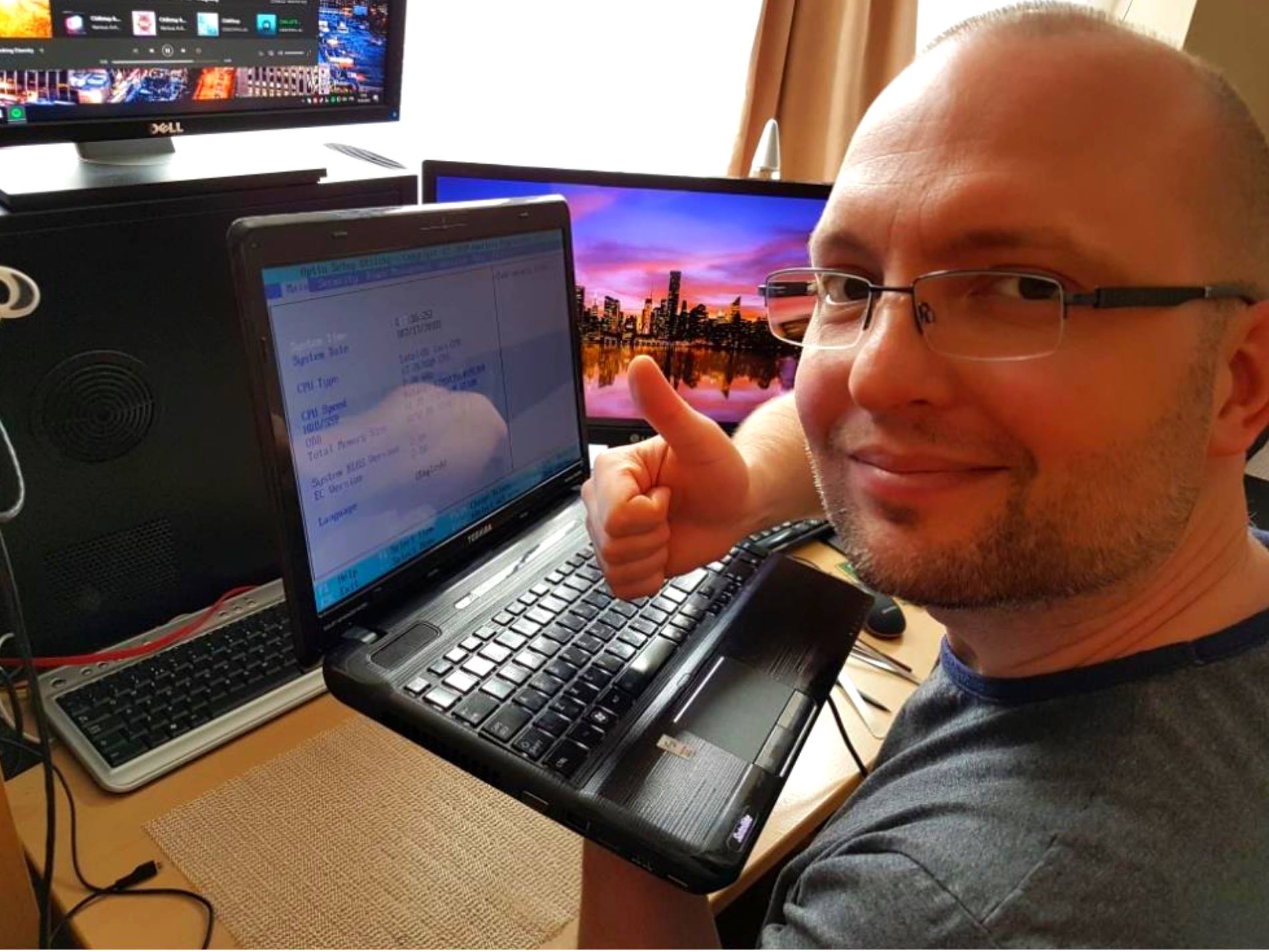 Uśmiechnięty mężczyzna w okularach trzyma laptopa Toshiba z ekranem BIOS, kciuk w górę, na biurku komputer stacjonarny i dwa monitory.