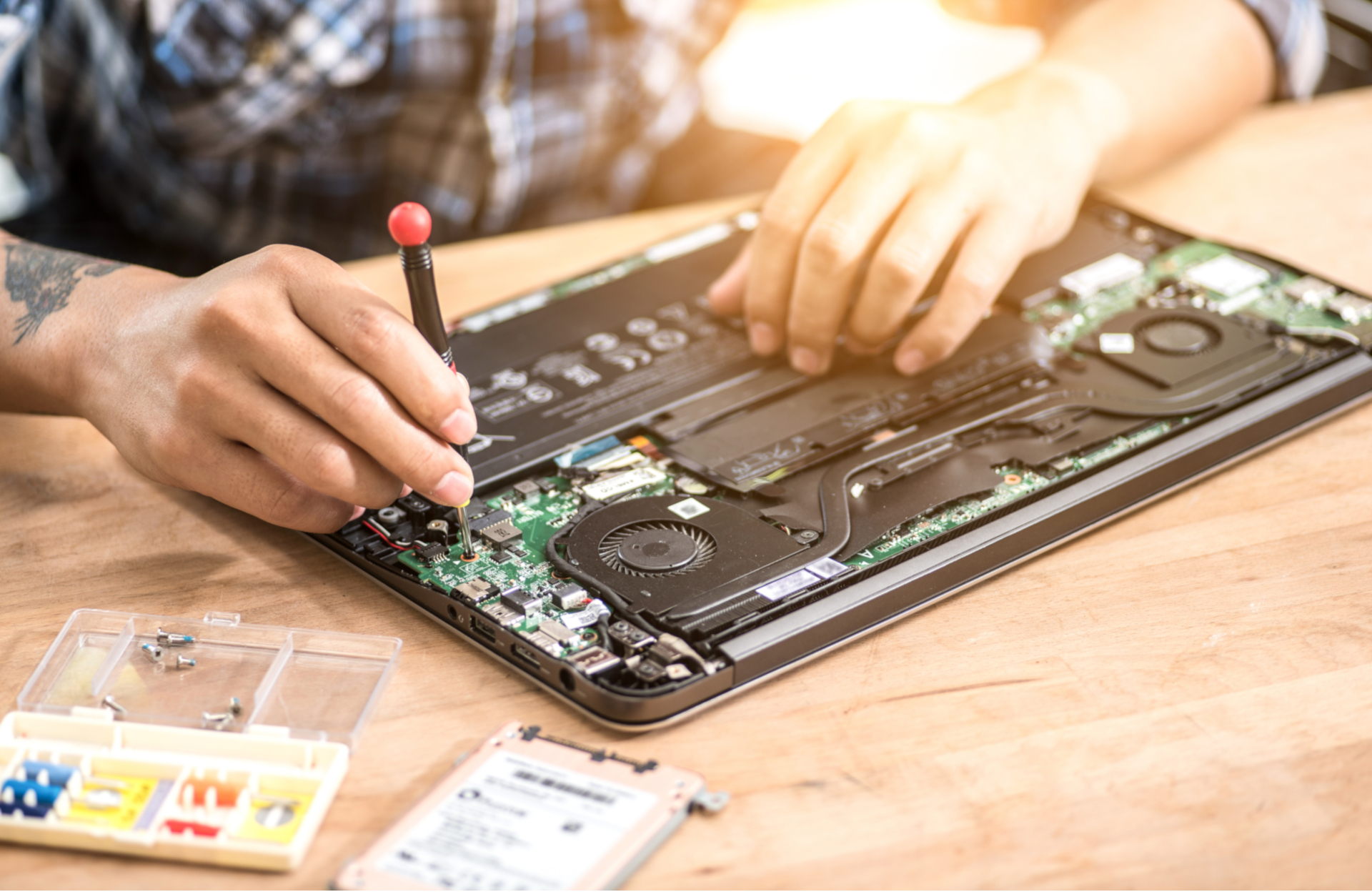Otwarty laptop na drewnianym stole, serwisant z tatuażem na ręce używa śrubokręta do naprawy płyty głównej, obok pudełko z elementami i dysk twardy.