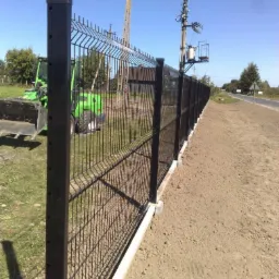 Długie, czarne ogrodzenie z siatki z betonową podmur&oacute;wką wzdłuż drogi, z zielonym traktorem rolniczym w tle i słupami energetycznymi na horyzoncie.