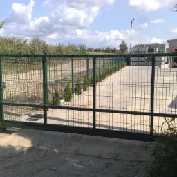 Przesuwna brama z zielonej siatki ogrodzeniowej, widok na teren z drzewami i budynkiem gospodarczym w tle.