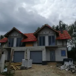 Surowy stan domu dwukondygnacyjnego z dwoma garażami w trakcie budowy, widoczne bloczki betonowe, częściowo położona dachówka i szare okna, materiały budowlane na placu budowy.