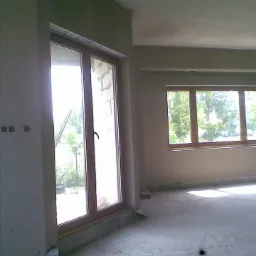 Wnętrze pomieszczenia w trakcie prac wykończeniowych, widoczne okna z drewnianymi ramami, ściany pokryte świeżym tynkiem, surowa podłoga w trakcie przygotowania.
