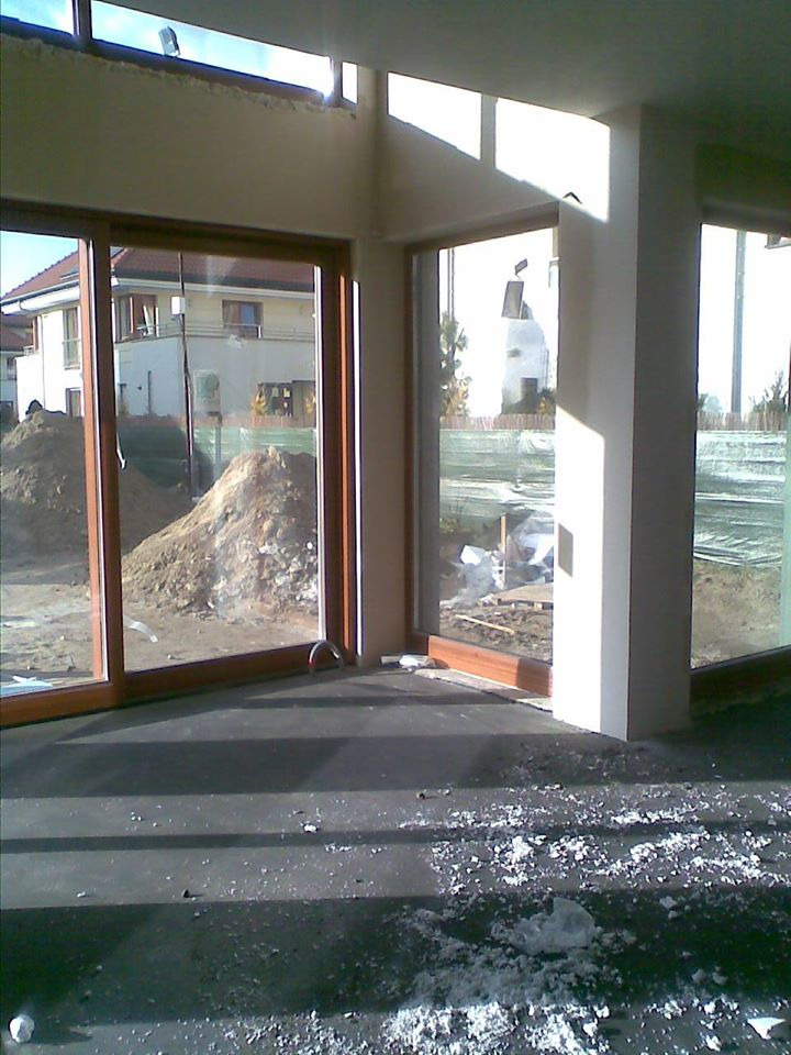 Wnętrze w trakcie prac wykończeniowych; widoczne duże okna z drewnianymi ramami, surowy tynk na ścianach i kolumnie, resztki styropianu na podłodze, za oknami hałda ziemi.