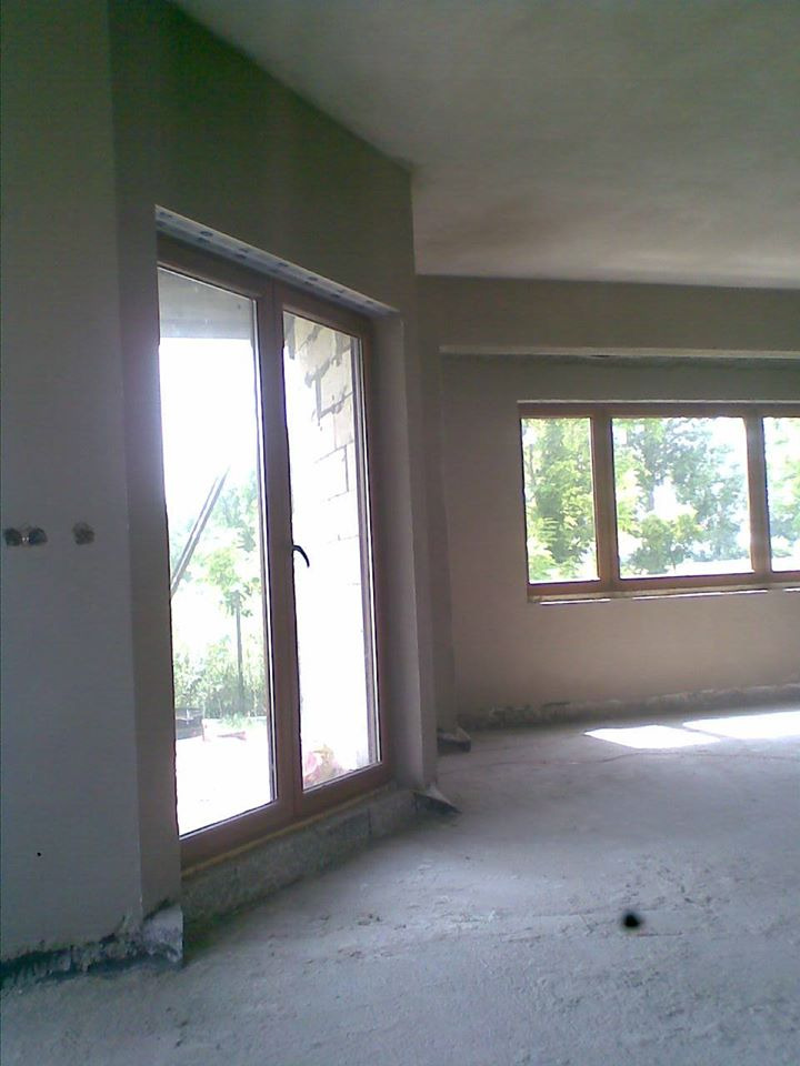 Wnętrze pomieszczenia w trakcie prac wykończeniowych, widoczne okna z drewnianymi ramami, ściany pokryte świeżym tynkiem, surowa podłoga w trakcie przygotowania.