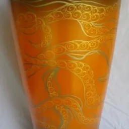 Wysoka, smukła waza z gradientem pomarańczowo-czarnym, zdobiona złotymi, spiralnymi ornamentami przypominającymi macki ośmiornicy, ustawiona na białym materiale.