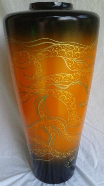 Wysoka, smukła waza z gradientem pomarańczowo-czarnym, zdobiona złotymi, spiralnymi ornamentami przypominającymi macki ośmiornicy, ustawiona na białym materiale.