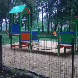 Nowoczesny plac zabaw z piaskownicą, otoczony czarnym ogrodzeniem panelowym w parku. Kolorowe elementy zabawowe na piaszczystym podłożu.