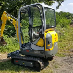 Żółto-szara mini koparka Wacker Neuson ET16 z otwartą kabiną, stojąca na trawiastym terenie obok drogi gruntowej z widocznymi śladami opon, w tle drzewa i częściowo wykarczowany teren.