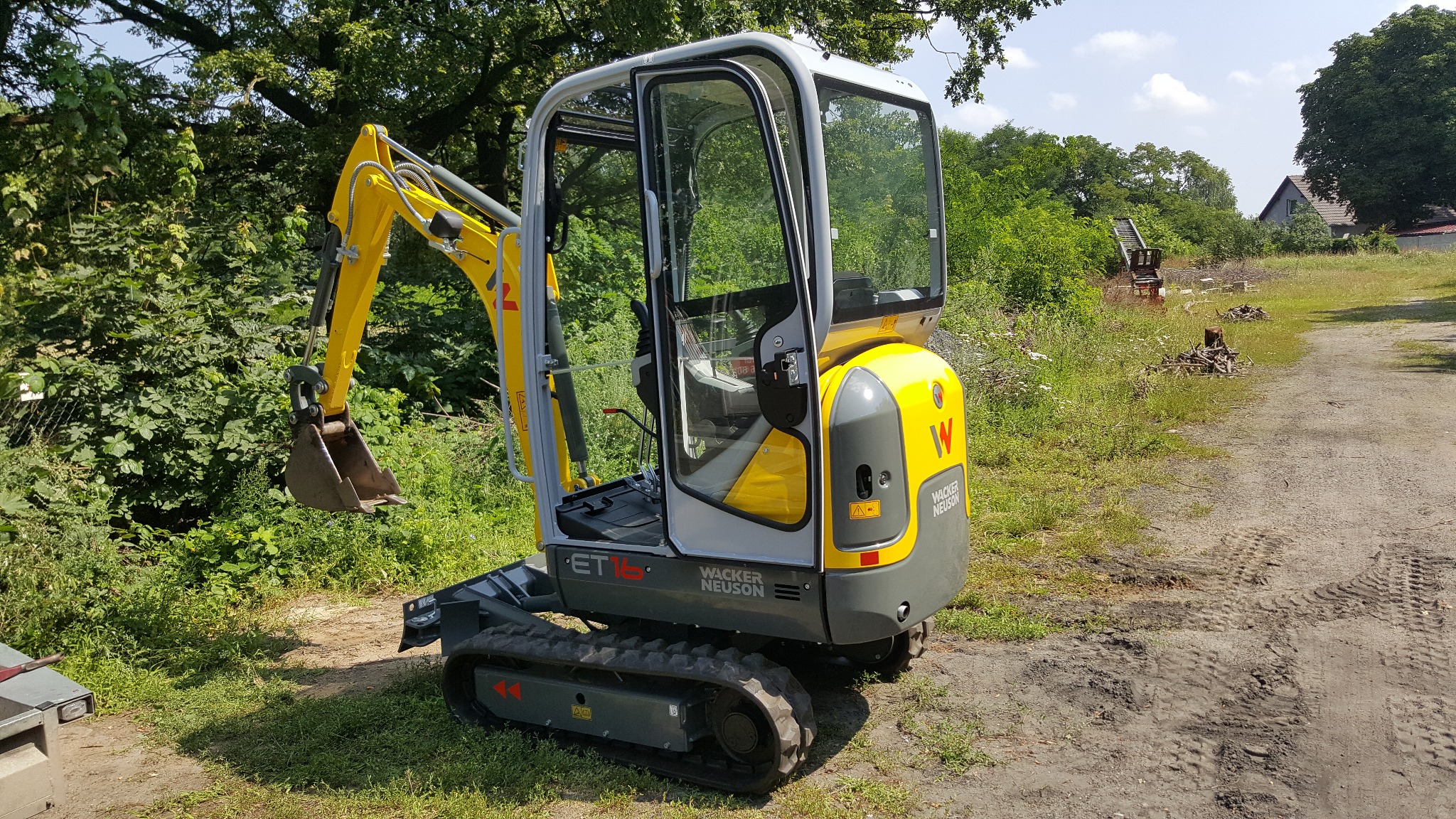 Żółto-szara mini koparka Wacker Neuson ET16 z otwartą kabiną, stojąca na trawiastym terenie obok drogi gruntowej z widocznymi śladami opon, w tle drzewa i częściowo wykarczowany teren.