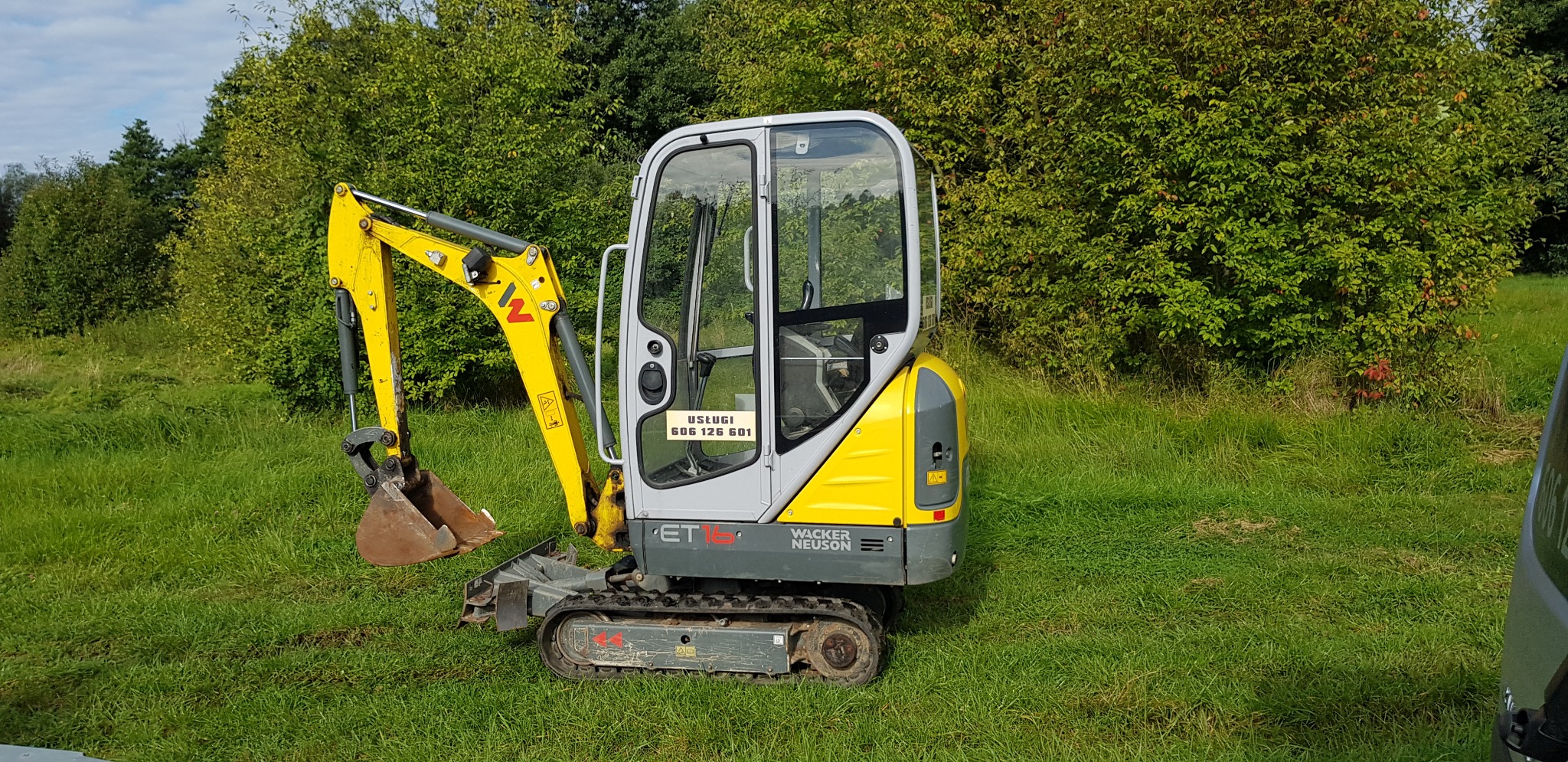 Żółto-szara mini koparka Wacker Neuson ET16 na gąsienicach, stojąca na trawiastym terenie z zielenią w tle, z naklejką z numerem telefonu na szybie kabiny.