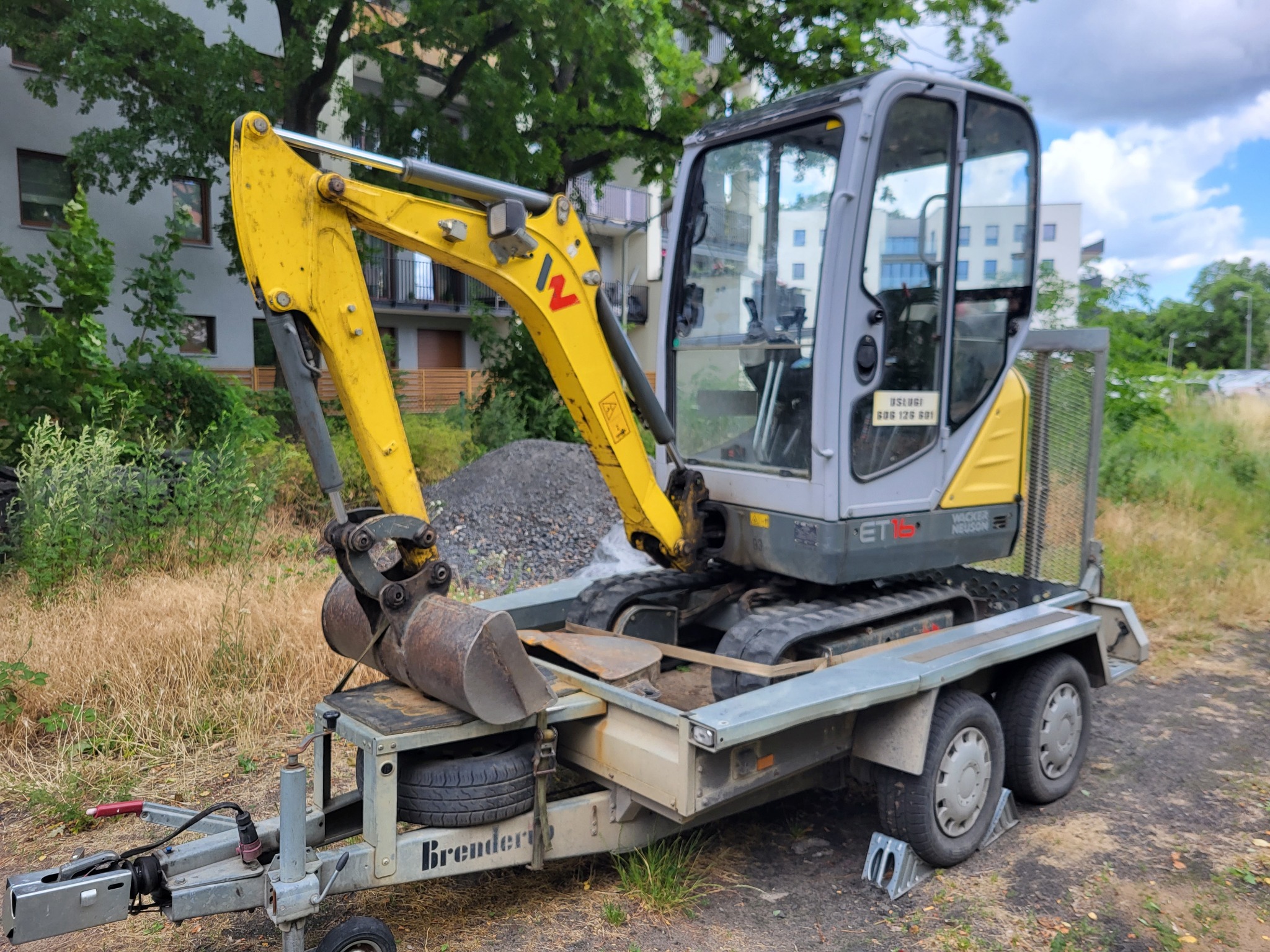 Żółto-szara mini koparka Wacker Neuson ET16 na trzyosiowej przyczepie Brenderup, zaparkowana na trawiastym terenie z budynkami w tle.