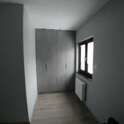 Szara szafa na wymiar z czterema drzwiami, dopasowana do ściany, z widocznym grzejnikiem pod oknem i drewnianą podłogą w minimalistycznym wnętrzu.