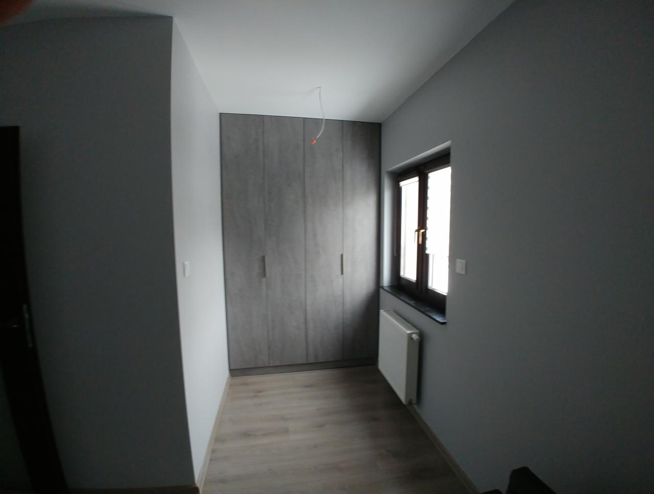 Szara szafa na wymiar z czterema drzwiami, dopasowana do ściany, z widocznym grzejnikiem pod oknem i drewnianą podłogą w minimalistycznym wnętrzu.