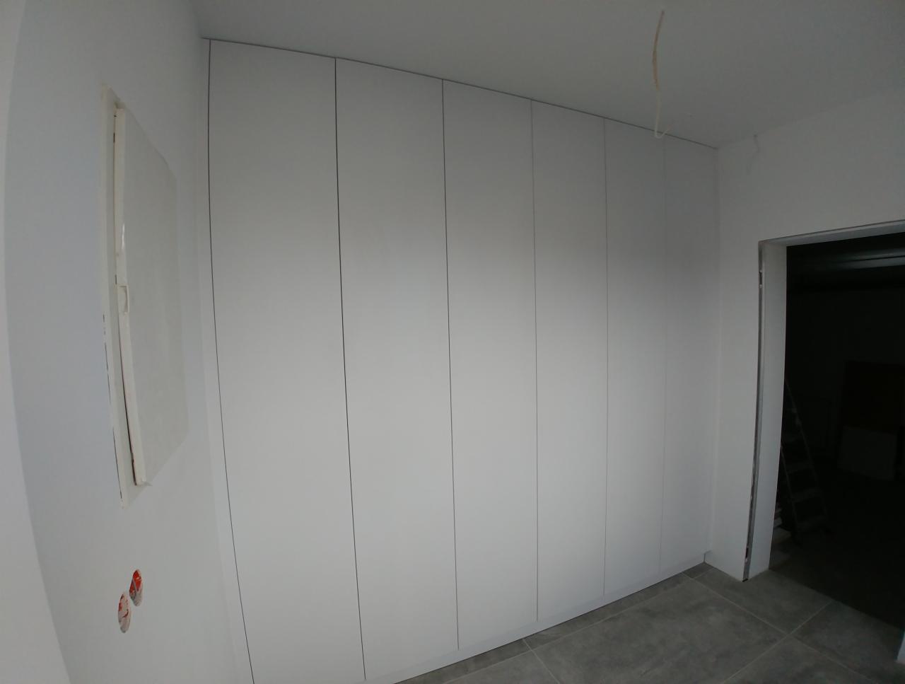 Biała zabudowa szafy wnękowej z drzwiami bez uchwytów, widoczna w surowym wnętrzu podczas remontu, szare płytki podłogowe.