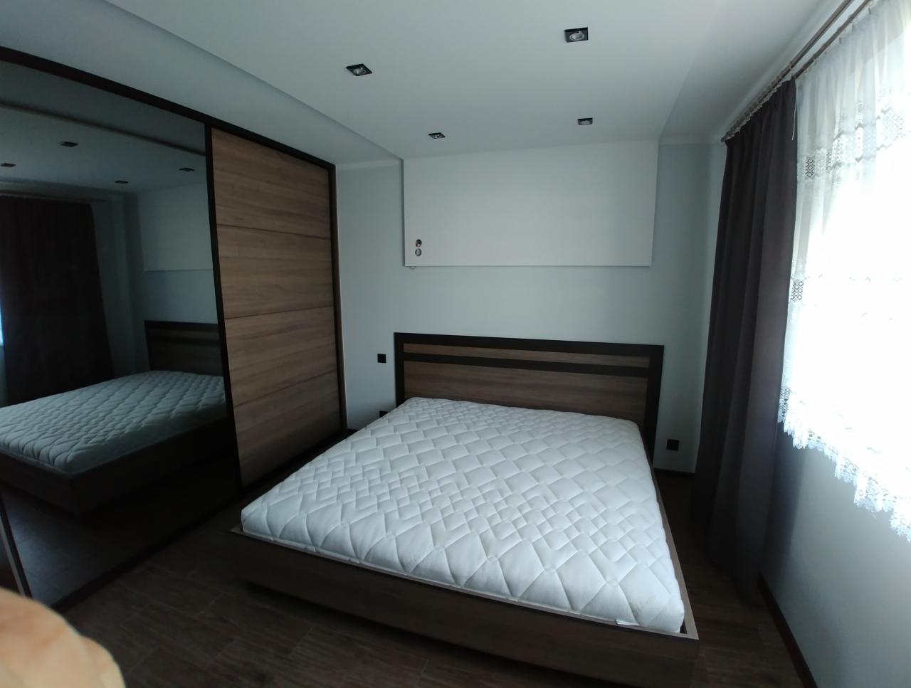 Sypialnia z łóżkiem i przesuwną szafą z lustrzanymi drzwiami, widoczne oświetlenie sufitowe i zasłony przy oknie.