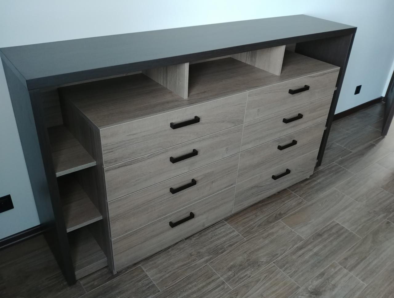 Szara komoda z szufladami i półkami, wykonana na wymiar, z ciemnymi uchwytami, ustawiona na podłodze z płytek imitujących drewno.
